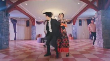 Geniş bir salonda, kareli zemini ve zarif dekoruyla dans eden bir grup insan. Dansçılar, geleneksel ve resmi kıyafetlerin karışımından giyinmiş, canlı bir atmosfer sergiliyorlar..