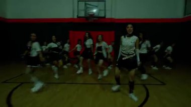 Bir grup dansçı şık kıyafetler giyerek bir spor salonunda gösteri yapıyorlar. Sahne, arka planda basketbol potası ile dinamik bir hareket ve enerji yakalıyor..