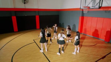 Bir basketbol salonunda dans rutini uygulayan şık kıyafetli altı genç kadın. Spor salonunda ahşap zemin, basketbol potası ve renkli duvar aksanları vardır..