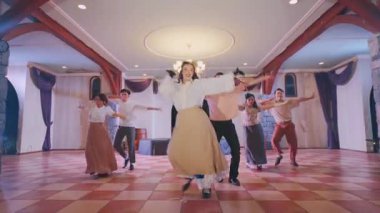 Bir grup dansçı, ahşap kirişler ve zarif dekorlarla geniş bir salonda canlı bir gösteri sergiliyorlar. Uzun etek ve eldiven giyen baş dansçı enerjik hareketler sergilerken, diğerleri koordineli adımlara katılıyor..