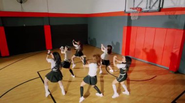 Bir spor salonunda sahne alan altı ponpon kızdan oluşan bir grup. Aynı kıyafetleri giyiyorlar, enerjik pozlar veriyorlar. Basketbol sahası arka planda, halka ve renkli duvarlarla görülebilir..
