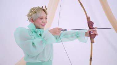 Nişan alırken gülümseyen, ok ve yaylı, nane yeşili kıyafetli biri. Çiçekli bir tacı ve neşeli bir ifadesi var. Yumuşak, tarafsız bir arkaplanı var..