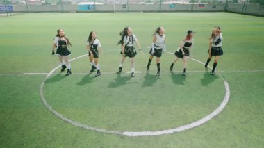 Futbol sahasında dans eden altı genç kadından oluşan bir grup. Aynı kıyafetleri giyiyorlar, enerjik hareketler sergiliyorlar ve gülümsüyorlar. Arkaplanda yeşil bir alan ve açık bir gökyüzü var..