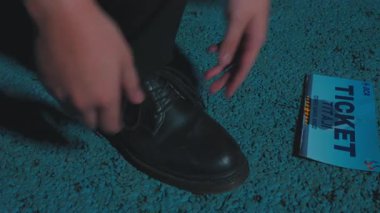 Ayakkabı bağcıklarını mavi halıya bağlayan birinin yakın çekimi. Odak noktası eller ve ayakkabılar. Arka planda kısmen görünür bir kitap var..