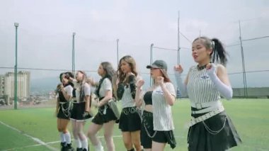 Üç dansçı spor sahasında enerjik bir performans sergiliyor, dinamik hareketler ve şık kıyafetler sergiliyor. Arka planda bulutlu bir gökyüzünün altında bir şehir manzarası var.