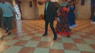 Geniş bir salonda kareli zemini olan canlı bir dans sahnesi. Birkaç çift dans ediyor, geleneksel kıyafetlerini sergiliyorlar. Yumuşak ışıklandırma şenlikli atmosferi güçlendirir.