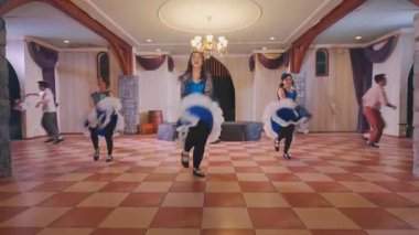 Canlı bir dans gösterisi. Mavi elbiseli, beyaz aksanlı üç kadın ahşap kirişler ve zarif dekorlarla neşeyle dans ediyorlar..