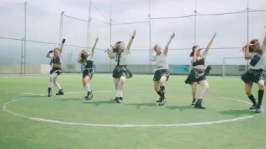 Altı dansçıdan oluşan bir grup yeşil bir çim sahasında koreografili bir gösteri yapıyor. Şık kıyafetler giyiyorlar, parlak bir gökyüzü altında enerjik hareketler sergiliyorlar..