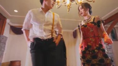 Güzel dekore edilmiş bir odada süslü bir avizenin altında dans eden neşeli bir çift. Kadın zarif, renkli bir elbise giyerken, erkek askılı sıradan bir beyaz gömlek giyer. İfadeleri mutluluk ve heyecan veriyor..