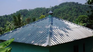 Açık mavi bir gökyüzünün altında yemyeşil ve tepelerle çevrili yeşil bir evin kıvrımlı metal çatısının yakın görüntüsü..