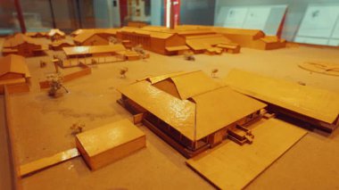 Geleneksel bir köy ya da yerleşim yerinin detaylı bir modeli, kumlu bir üste sergilenen ahşap yapılar ve binalar. Modelde, muhtemelen tarihi veya kültürel bir alanı temsil eden çeşitli binalar yer alıyor..