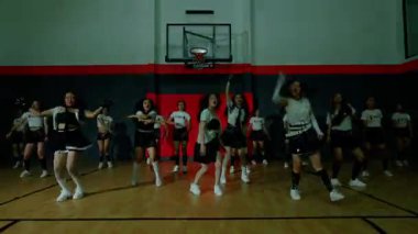 Bir grup dansçı jimnastik salonunda senkronize hareketler sergiliyor. Birbirlerine uyumlu kıyafetler giyiyorlar. Sportif bir havaları var. Güce ve takım çalışmasına önem veriyorlar..