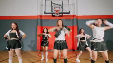 Bir spor salonundaki dinamik dans gösterisi, şık kıyafetler giymiş iki dansçının yer aldığı. Bir dansçı odak noktasında, enerjik hareketler sergiliyor, diğeri ise arka planda biraz bulanık. Set bir basketbol potası ve renkli bir çember içeriyor.