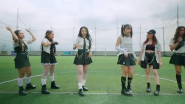 Çimenli bir alanda senkronize dans eden altı genç kadından oluşan bir grup. Şık kıyafetler giyiyorlar, etek ve botlar giyiyorlar, spor arkaplanı ve bulutlu gökyüzü var..