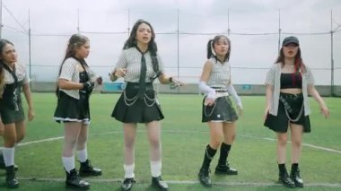 Bir spor sahasında enerjik bir şekilde dans eden, şık kıyafetler giyen beş genç kadından oluşan bir grup. Sahne hareketli ve dinamik bir atmosfer yakalıyor. Bulanık hareket, hareketi gösteriyor..