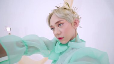 Kısa, açık renkli saçlı, nane yeşili elbisesi ve zarif süsleri olan birinin yakın çekimi. Ellerinde bir makas, yumuşak ve manevi bir arka planda durarak poz veriyorlar..