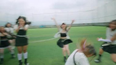 Çitlerle çevrili çimenli bir alanda dans eden beş genç kadından oluşan bir grup. Şık kıyafetler giyiyorlar, enerjik hareketler ve ifadeler sergiliyorlar. Gökyüzü bulutlu, sahneye dramatik bir zemin ekliyor.