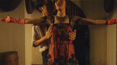 Klasik bir ortamda bir erkek ve bir kadının olduğu dramatik bir sahne. Erkek, sıkıntılı görünen kadının hemen arkasında duruyor. Kadın süslü bir elbise ve mücevher giyer, erkek ise resmi giyinir. Işık gergin bir atmosfer yaratıyor..