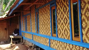 Mavi ahşap süsleme ve geniş pencereli dekoratif bambu kaplamalı geleneksel bir ev. Yapı, arka planda ağaçlar olan doğal bir ortamda kurulmuştur..