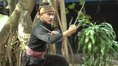 Geleneksel kıyafetli bir dövüş sanatçısı doğal bir ortamda silahla poz verir. Dekoratif kemeri ve saç bandı olan siyah bir kıyafet giyiyor..