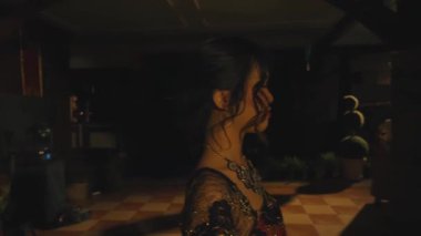 Koyu renk saçlı, kıvırcık saçlı, şık bir kıyafet ve bir kolye takan bir kadının yan profili. Arka plan dekoratif elementlerle loş bir iç mekan içeriyor.