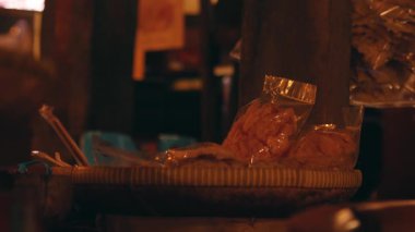 Örgü sepetine konmuş poşetlerle dolu loş bir pazar sahnesi. Atmosfer sıcak ve davetkar. Atıştırmalıklara odaklı..