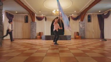 Beş dansçıdan oluşan bir grup, ahşap kirişleri ve zarif dekorlarıyla geniş bir salonda gösteri yapıyorlar. Dansçılar renkli kostümler giyer, canlı bir dans gösterisi yaparlar. Atmosfer neşeli ve enerjik..