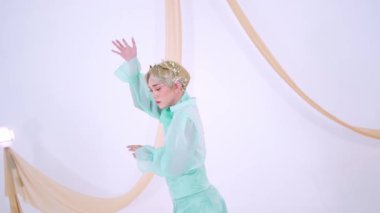 Nane yeşili kıyafetli bir kişi, akıcı kumaşlı minimalist bir geçmişe karşı zarif bir şekilde poz verir. Elleri zarif bir şekilde kaldırılır, zarif bir stil sergilerler.