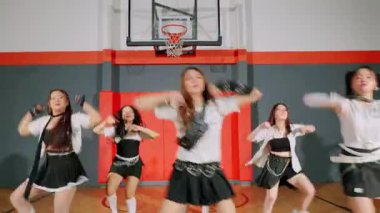 Bir grup dansçı bir spor salonunda sahne alıyor, siyah etekli ve beyaz bluzlu şık kıyafetler giyiyorlar. Dinamik pozisyondalar, koreografilerini sergiliyorlar..