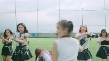 Bir grup dansçı spor sahasında gösteri yapıyor, dinamik hareketler sergiliyorlar. Ön planda oyuncu bir ifadeye sahip bir dansçı yer alırken, diğerleri onun arkasında hareket halindedir. Ayar parlak ve enerjik, dans gösterisi için ideal..