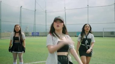 Bulutlu bir gökyüzünün altındaki bir spor sahasında dans eden üç genç kadın. Şık kıyafetler giyiyorlar. Şapkalar, spor giysileri, enerji ve coşku gösterisi..