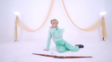 Zarif bir şahıs yerde zarif bir şekilde poz verir, nane yeşili bir kıyafet giyer. Arka planda yumuşak perdeler ve ışık var, sakin bir atmosfer yaratıyor. Önünde bir okçuluk yayı var..