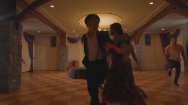 Bir grup dansçı, loş bir balo salonunda geleneksel kıyafetlerini sergilediler. Odak noktası ön planda dans eden bir çift ve arka planda hareket eden diğerleri..