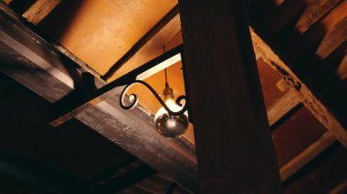 Kırsal bir ortamda ahşap bir kirişe asılmış klasik tarzda bir lamba loş bir odada sıcak bir ışık saçıyor..