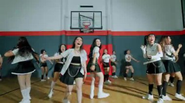 Basketbol salonunda gösteri yapan bir grup genç dansçı. Şık, sportif kıyafetler giyiyorlar ve koreografili bir dans gösterisi sergiliyorlar. Atmosfer enerjik ve enerjik. Arka planda basketbol potası var..