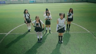 Bir spor sahasında dans eden altı genç kadından oluşan bir grup. Şık kıyafetler giyiyorlar, canlı yeşil çimen arka planlı, etek ve üstlerin karışımını sergiliyorlar..