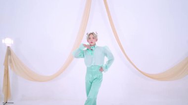 Tarz sahibi bir birey açık yeşil bir elbisenin içinde, katıksız bir üst ve yüksek bel pantolon ile güven içinde poz verir. Arka plan yumuşak perdelerle minimalistik, zarif bir atmosfer yaratıyor..