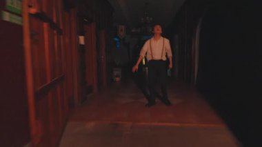 Beyaz gömlekli ve pantolon askısı takmış bir adam loş bir koridorda duruyor ve şaşırmış bir ifadeyle yukarı bakıyor. Koridorda birkaç dekoratif unsurla ahşap duvarlar ve kapılar var..