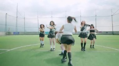 Altı dansçıdan oluşan bir grup yeşil bir çim sahasında, şık siyah beyaz kıyafetler giymişler. Dinamik pozlarda kareografilerini bulutlu bir gökyüzü altında sergiliyorlar..