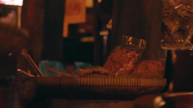 İçinde paketlenmiş atıştırmalıklar olan dokuma sepetinin olduğu loş bir sahne, kırsal bir arka plana ayarlanmış. Atmosfer sıcak ve davetkar, yerel lezzetleri sergiliyor..