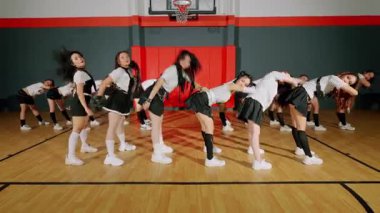 Bir grup genç kadın spor salonunda senkronize dans gösterisi yapıyor. Çizgili üst ve etekleri olan uyumlu kıyafetler giyiyorlar, enerjik hareketler sergiliyorlar. Arka plandaki basketbol potası sportif atmosfere katkı sağlıyor..