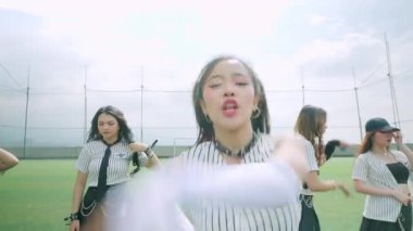 Bulutlu bir gökyüzünün altında çimenli bir alanda dans eden bir grup genç kadın. Şık kıyafetler giyiyorlar, gündelik ve keskin elementlerin karışımı, enerjik hareketler sergiliyorlar..