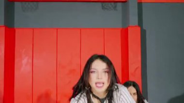 Kırmızı ve gri arkaplanlı canlı bir ortamda enerjik bir performans sergileyen iki dansçı. Bir dansçı etkileyici bir pozla hareket ederken, diğeri daha rahat ve dans kıyafetlerini sergiliyor..