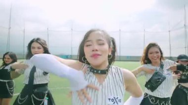 Bir grup genç dansçının çimenli bir alanda performans sergilediği canlı bir sahne. Ön planda, çizgili bir kıyafet giymiş, at kuyruklu bir kıza odaklanıyoruz. Arka plan diğer dansçıları ve bulutlu gökyüzünü gösteriyor..