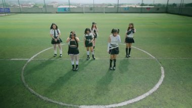 Futbol sahasında dans eden altı genç kadından oluşan bir grup. Şık kıyafetler giyiyorlar, etekleri ve koordine edilmiş üstleri var, canlı yeşil çim arka planı var..