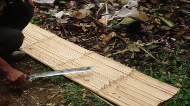 Tahta bir tahtanın üzerinde çalışan bir insan, onu oymak ya da şekillendirmek için bir araç kullanır. Arka planda yapraklar ve molozlar olan dağınık bir alan var. Bu da açık hava ortamını gösteriyor..