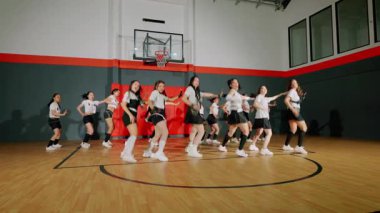 Bir grup dansçı bir spor salonunda koreografi gösterisi yapıyor. Şık kıyafetler giyiyorlar ve enerjik hareketler sergiliyorlar. Arka planda basketbol potası ve ahşap zemin var..