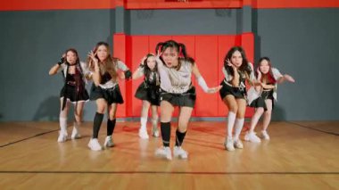 Dinamik bir dans gösterisi, şık kıyafetler içindeki beş genç kadından oluşan bir grup, kırmızı ve gri duvarlı bir spor salonu ortamında enerjik hareketler sergiliyor..
