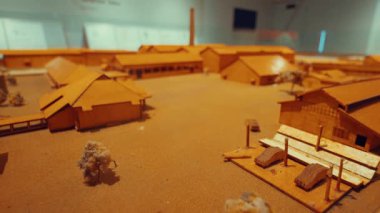 Ahşap binalar, ağaçlar ve araçların bulunduğu tarihi bir köy veya sanayi bölgesinin detaylı bir mimari modeli, hepsi kumlu bir zemine kurulmuş. Model, karmaşık tasarım ve düzeni sergiliyor..