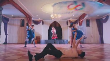 Bir grup dansçı ahşap kirişleri ve kareli zemini olan geniş bir salonda gösteri yapıyorlar. Bir dansçı ön planda, mavi bir kumaşı sallarken diğerleri arka planda dans ediyor..